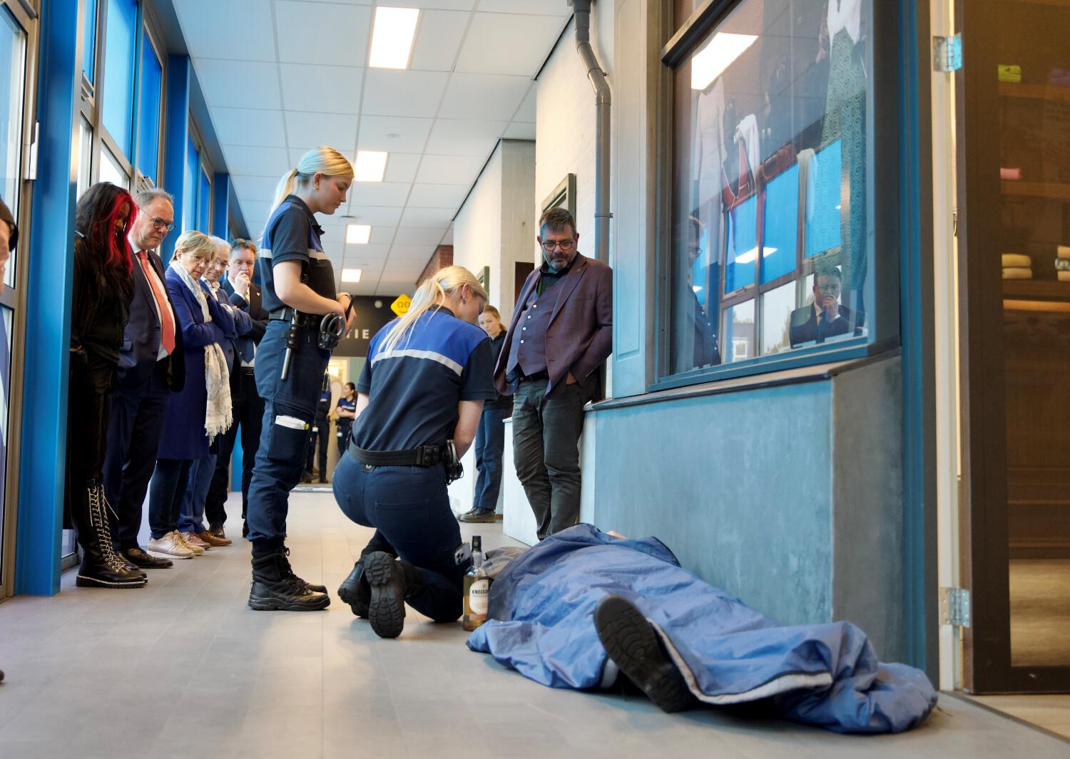 Twee blonde Aventus-studentes in uniform buigen zich in een praktijkoefening over iemand die onder een deken op de gang ligt met een fles sterke drank naast zich