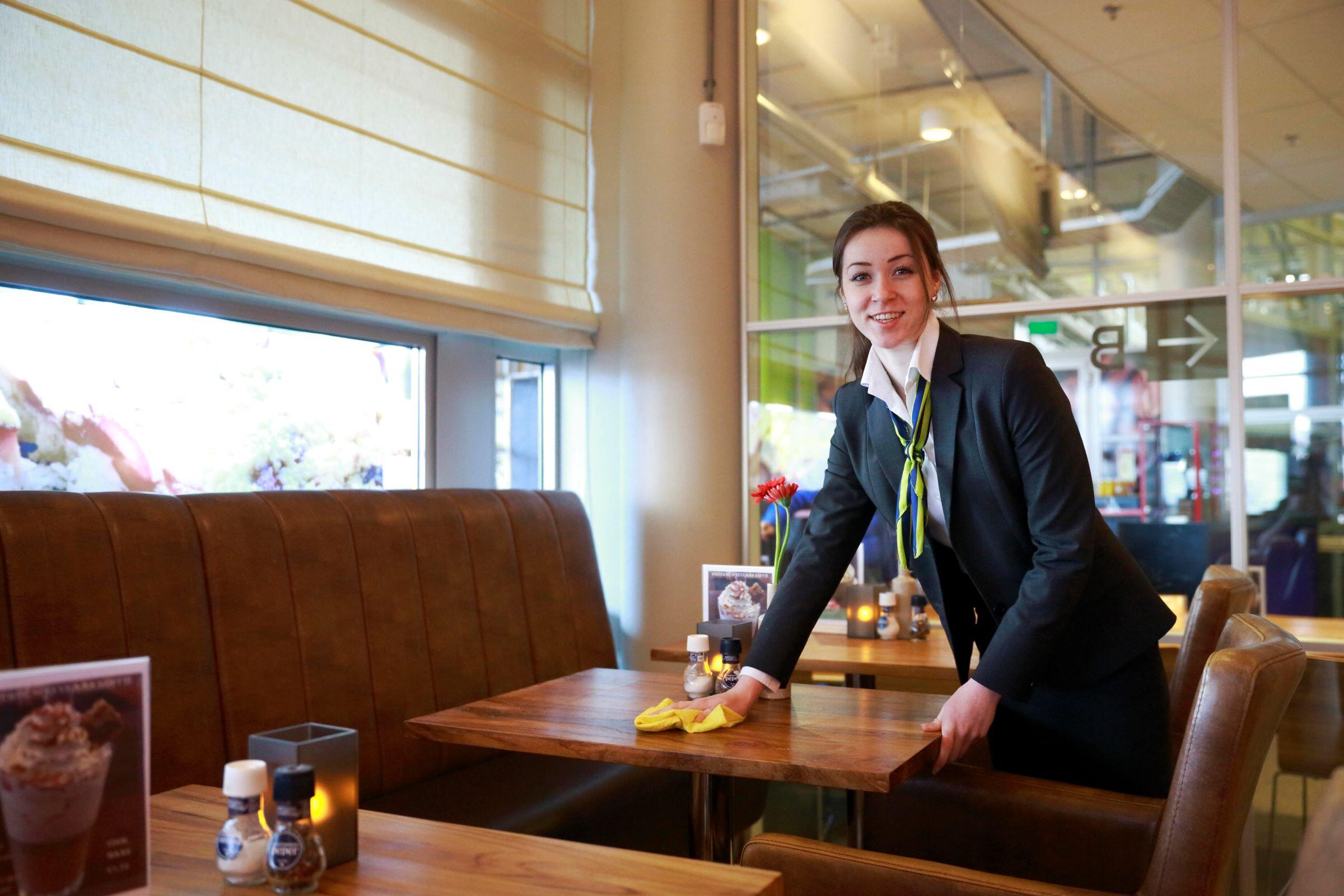 Een Aventus-studente Horeca kijkt vriendelijk in de camera en maakt de tafel in de brasserie schoon met een doekje