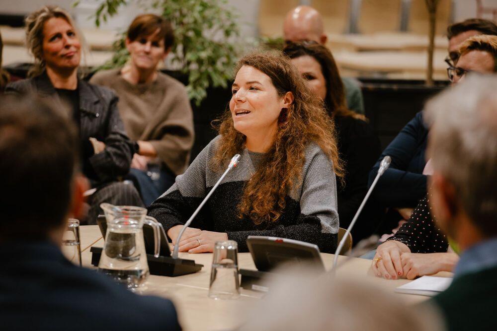 Een dame zit aan een tafel en praat in een microfoon, op de achtergrond zit het publiek