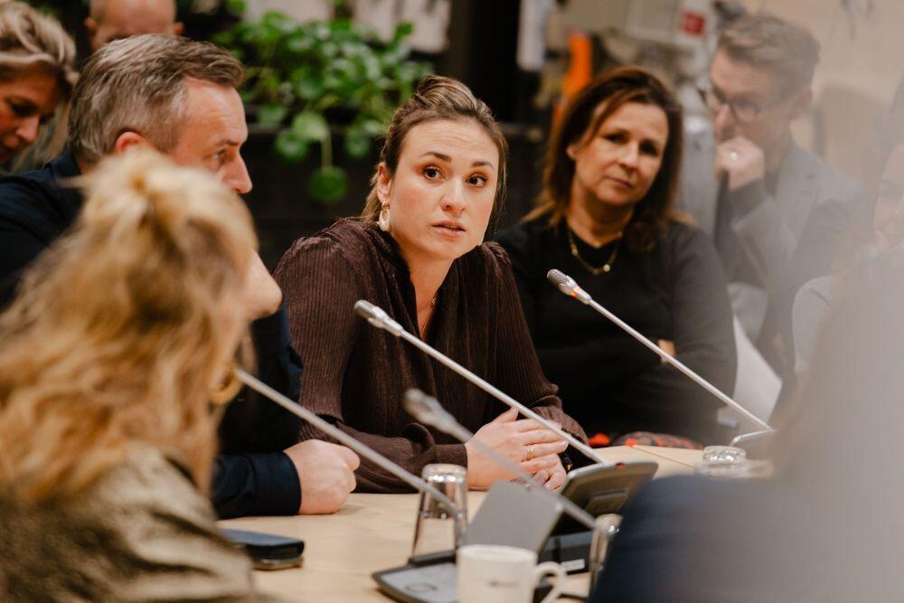 Een aantal mensen zit  te luisteren aan een tafel met microfoons op de tafel