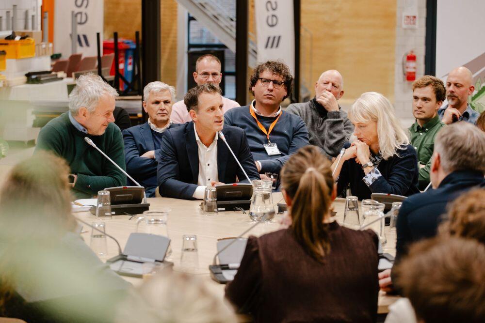 Een aantal mensen zit  te luisteren aan een tafel met microfoons op de tafel