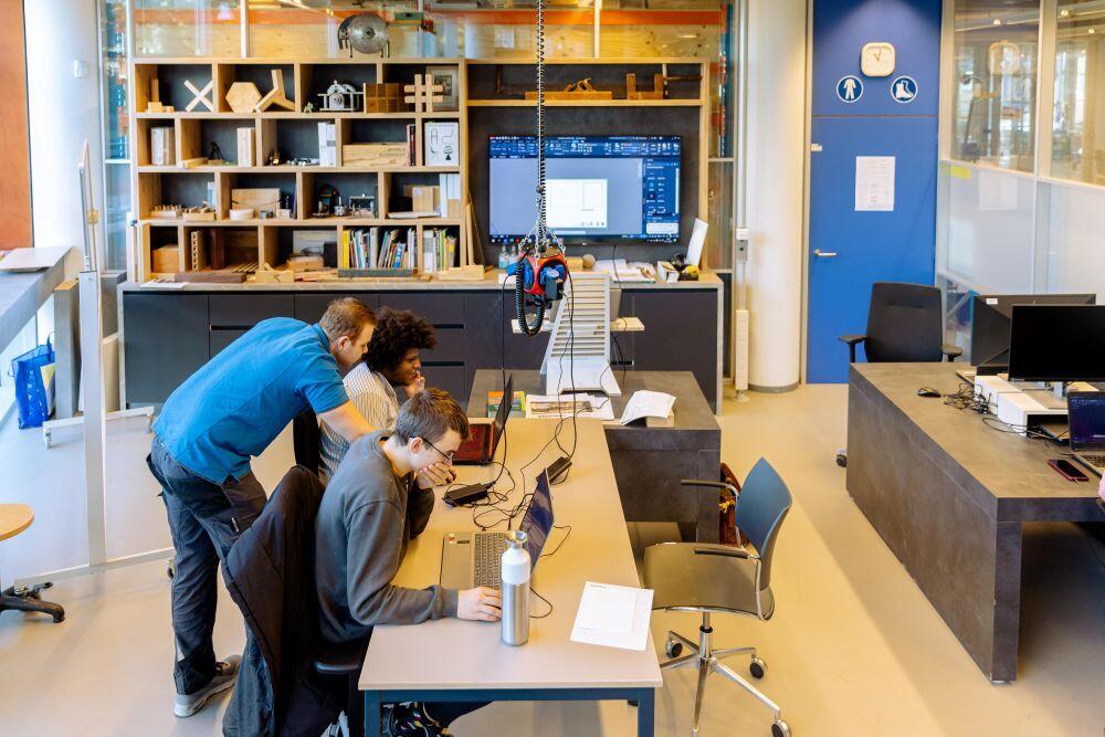 Twee studenten Hout & Meubel zitten aan een tafel achter een laptop in de praktijkruimte en een docent helpt hen