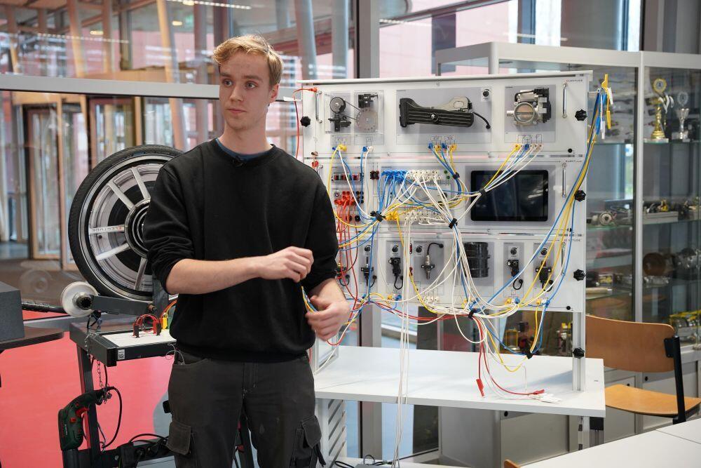 Een jongeman staat in de praktijkruimte van Autotechniek voor een paneel met autolampen etc. en houdt een bundel elekticiteitsraden vast