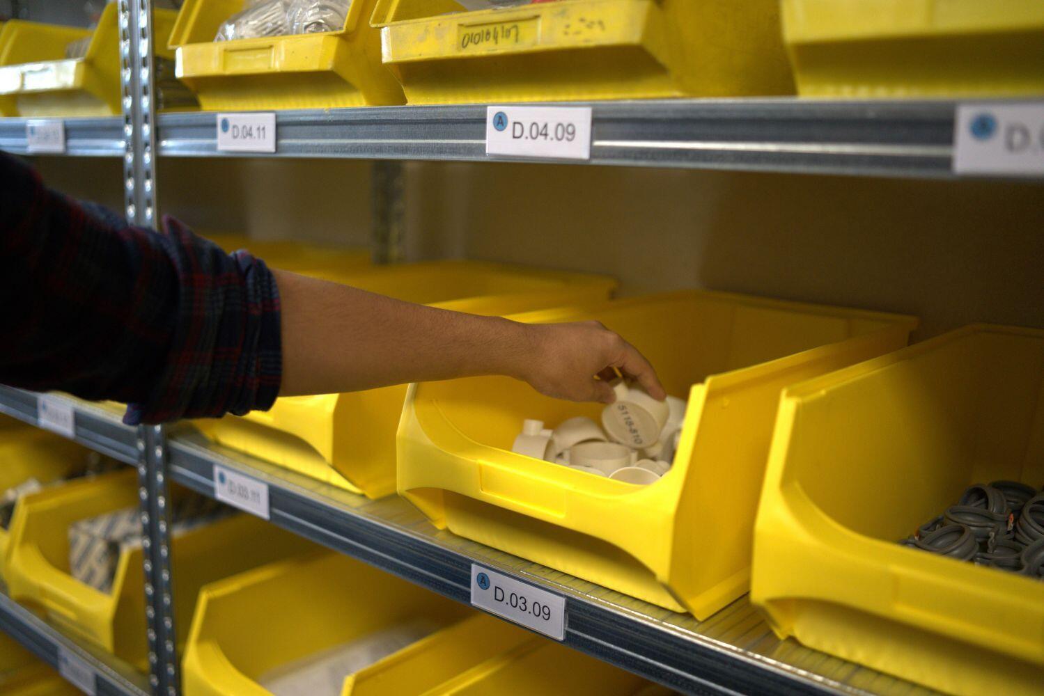 Een hand pakt iets uit een gele bak in een magazijn