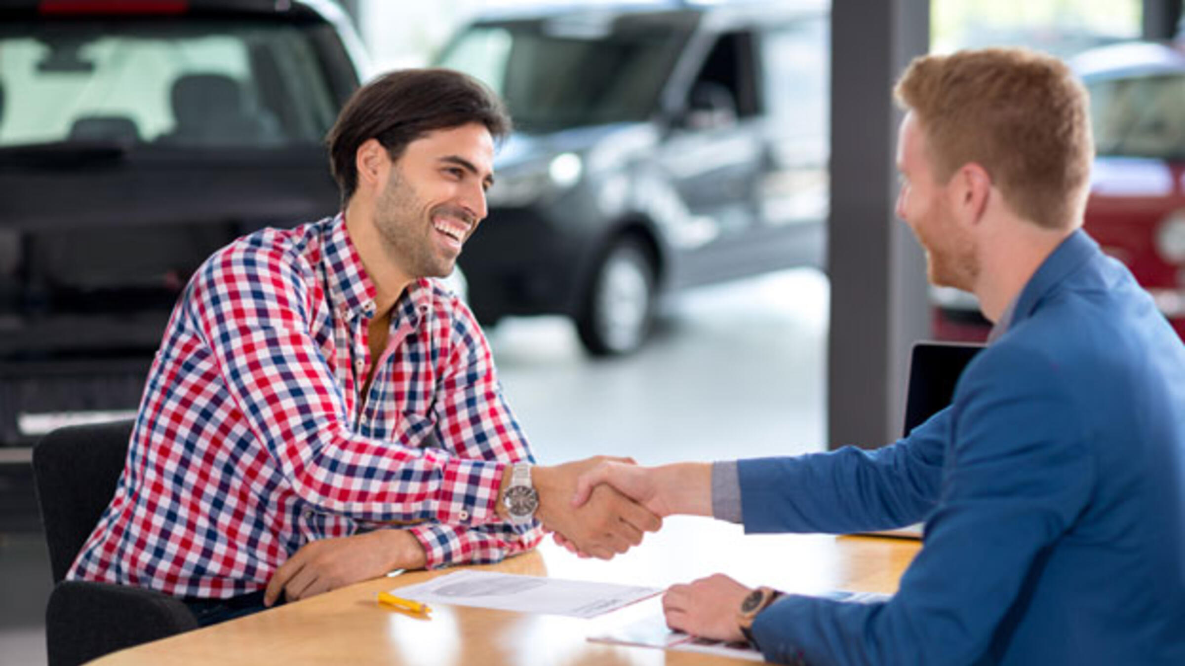 Twee mannen zitten aan  een tafel bij een autodealer en ze schudden elkaar lachend de hand